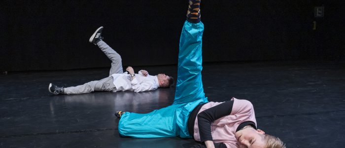 Anno und Lukas liegen auf dem schwarzen Tanzboden, Anno vorne rechts, Lukas hinten links. Ein Bein ist jeweils senkrecht nach oben gestreckt.