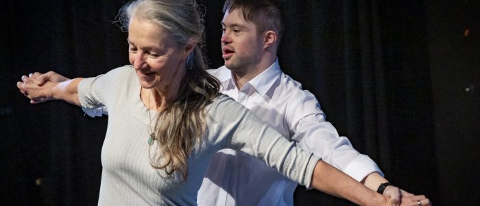 Sarah und Lukas stehen eng hintereinander, die Arme zur Seite ausgestreckt, sie fassen sich an den Händen. Eine vertraute und nahe Pose von Mutter und Sohn. Sie lächeln.