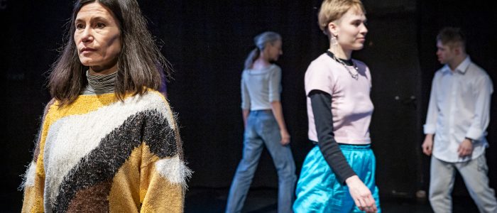Die Performer*innen gehen auf der Bühne herum. Vorne links Keiko in einem gelb-gemusterten Strickpullover, rechts Anno mit einem rosa T-Shirt und in türkiser Sporthose. Im Hintergrund Sarah und Lukas in weiß-grau.