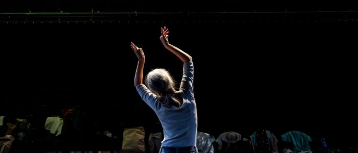 Sarah steht vor den leeren Zuschauerreihen, ihr Körper ist von vorne beleuchtet, die Arme sind filigran nach oben gestreckt, die Finger gespreizt.