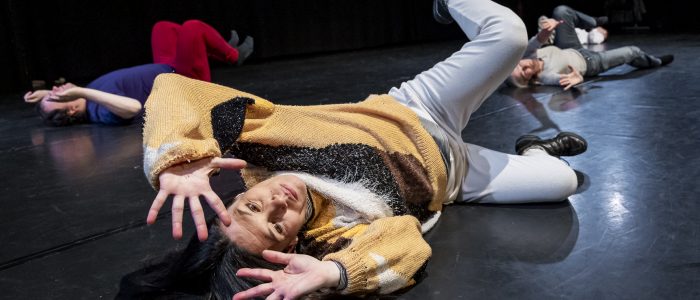 Die Performer*innen liegen mit dem Rücken auf dem schwarzen Tanzboden, Arme und Beine in die Luft abgewinkelt. Die Körper formen eine Art Kugel.