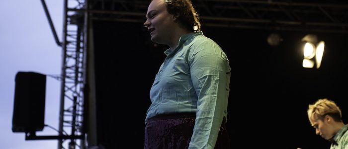 Lilian steht auf der Bühne der Night of Dance und spricht zum Publikum. Dey blinde Performer*in trägt eine türkise Bluse und einen bordeauxfarbenen Paillettenrock.