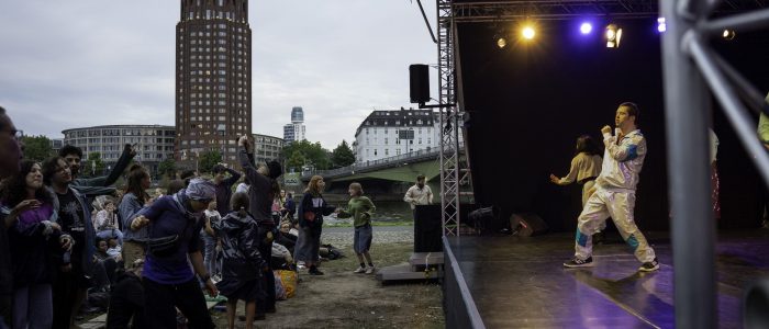 Blau-gelbes Bühnenlicht, Lukas in schimmerndem weiß-türkisen Sportanzug singt zu Queen: Don't stop me now. Vor der Bühne viele Menschen, die tanzen, im Hintergrund der Main und der Main Plaza Turm in Sachsenhausen.