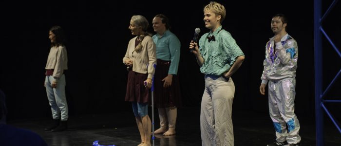 Alle fünf Performer*innen stehen gleichmäßig verteilt auf der Bühne der Night of Dance, Anno hält ein Mikrofon in der Hand, Sarah eine Krücke, um sich zu stabilisieren, die andere am Boden.